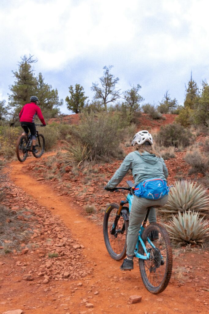 Two cyclists navigate a rugged mountain trail under a cloudy sky.